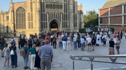 Lots of people are standing on concrete outside a church. Most of them have their backs towards the camera and are looking towards the entrance of the church, where a choir is singing.