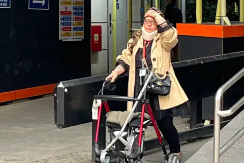 PA Media Woman has left hand on forehead while pushing a walker with her right hand. a crutch is sticking out