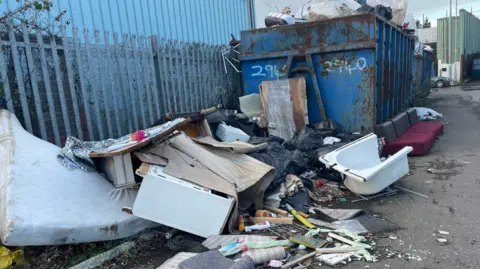 An image of rubbish spread out on the road. There is a bath and other items spread out across the floor. A huge, blue metal container stands behind them, next to a fence.