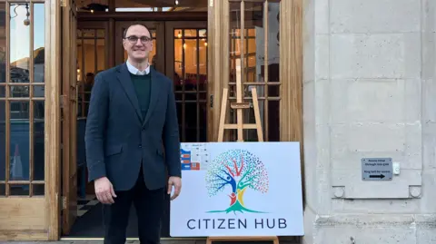 Harriet Heywood/BBC Ian Sollom standing outside the new hub next to a white sign which reads 'Citizen Hub'. He is wearing a suit, circle-framed glasses and a shirt