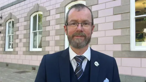 Deputy Malcolm Ferey is smiling at the camera as he stands on the pavement outside a pink granite building. He has short grey and brown hair and a beard and is wearing glasses. He is wearing a navy jacket and waistcoat over a white shirt and navy and pale blue striped tie. He has a silver pin badge and a pale blue handkerchief in his top pocket.