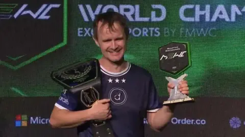 MEWC A man with short brown hair is smiling, holding a wrestling-style belt and a trophy. That's a navy-blue e-sports jersey with sponsors on the sleeves. Behind him is a display board with the Microsoft Excel World Championship and the event sponsors listed below.