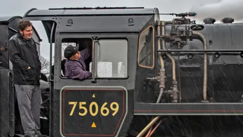 The footplate crew of  driver Mick Mathews and fireman Alex Robinson on a train on the heritage line. Mr Matthews is sitting in the carriage and Mr Robinson is standing on a ledge on the side of the train, which is black with gold lettering.