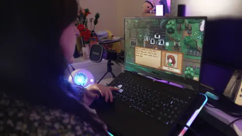 Sarah sat at a desk with her back to the camera, with her had on a gaming keyboard and a screen in front of her showing a computer game