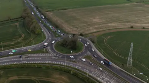 An aerial view of a large roundabout with a grassy central island and three exits. There are trees in the centre and multiple lanes coming off at each exit. The roundabout is set between grassy fields. Buildings of a residential area are int eh background.