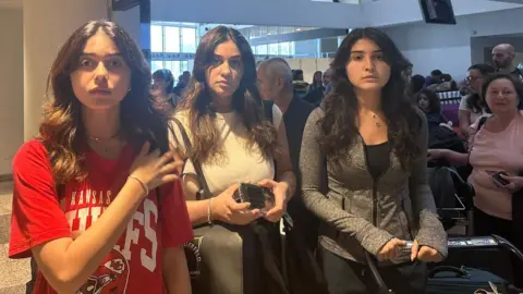 BBC Amal Zahereddine wearing a red t-shirt with her sisters Yasmine and Layla, all looking with sad expressions on their faces pictured in an airport terminal