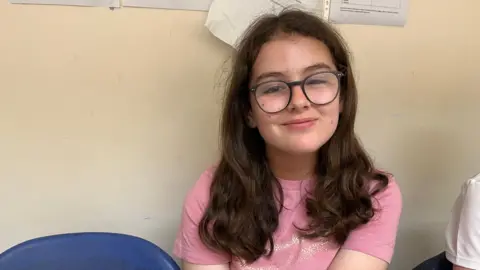 Ollie Conopo/BBC Freya has long dark-brown hair and glasses and is wearing a pink t-shirt. She is sitting on chair with a light-coloured wall behind her.