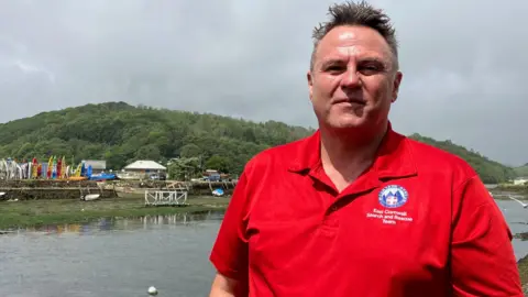 Dr Bruce Hughes is standing in front of a river. He is smiling and has brown hair which is short at the sides and longer on top. He is wearing a red polo shirt with East Cornwall Search and Rescue Team and a logo on the chest pocket. On the opposite bank is an array of paddle boards in storage.