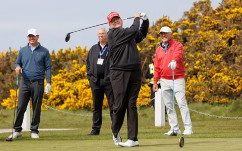Pa Trump jest za nim w swojej huśtawce golfowej, po prostu uderzył piłkę z kierowcą. Jest ubrany na czarno z białymi pokazami golfowymi i czerwoną czapką mag. Za nim są trzech mężczyzn, dwóch z nich wygląda, jakby grają z nim w golfa. Za nimi wszystko znajduje się zbiór krzewów w żółtym kwiacie