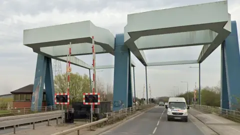 Google Twin blue and grey lifting bridges, which carry a dual carriageway over the River Hull. To the left are raised red and white barriers and warning lights, while to the right a white van travels towards the camera.