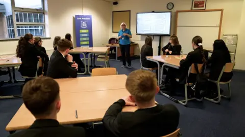 A classroom with 11 male and female pupils sitting at tables listen to a presentation from a woman wearing a blue polo shirt 