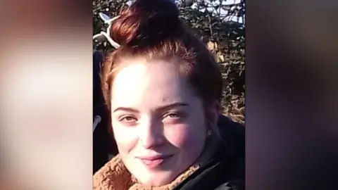 Family A young woman with brown hair in a top-knot is looking at the camera. She is wearing a coat with a fleece collar