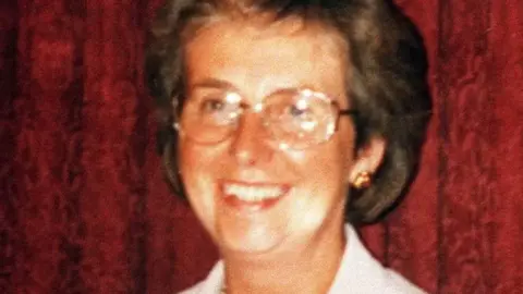 Family Handout Woman with short brown hair and large round glassed standing in front red curtain