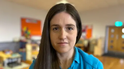 A woman with dark hair, wearing a blue top, and standing in a room which is out of focus in the background. 