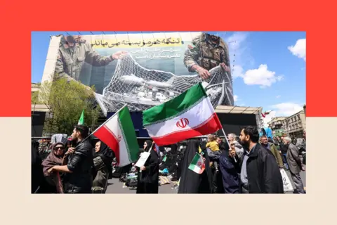 AFP via Getty Images Iranians hold national flags beneath a large billboard reading "The Strait of Hormuz remains closed" as people gather in Tehran's Revolution Square