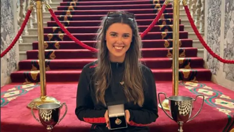 Bronagh Lennon  Méabh holding her medal. She's sitting on carpeted stairs with two sport cups beside her.