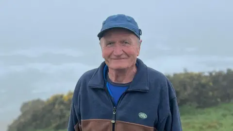 David Matthews is standing in a clifftop field in the fog with the sea and waves barely visible in the background. A gorse patch is also visible behind him. David is wearing a blue and brown zip up fleece with a Land Rover logo on the left side and a blue baseball cap.