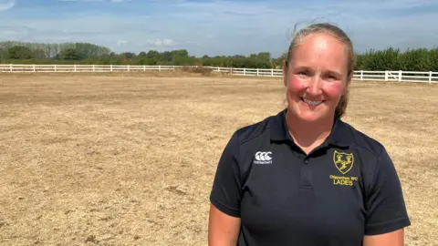 Clare Avenell stands on a dried out sports pitch, the grass is brown and yellow after weeks of rain. She is wearing a black polo top with a yellow club emblem on it and has blonde hair which is tied back