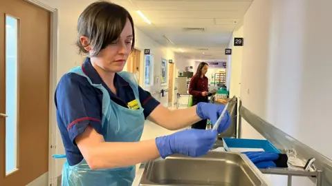 A nurse wearing a plastic apron and a uniform and blue gloves holding equipment near a sink. 