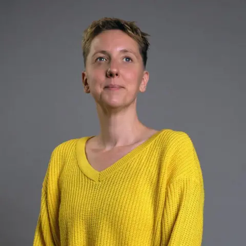 BBC Scotland A woman with short, cropped hair. She wearing a yellow V-necked jumper, and is sitting proud, looking into the distance.
