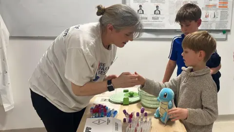 Richard Knights/BBC A medical staff member interacts with a young boy over a table which contains lots of vials. He holds a toy turtle in one hand while the lady puts something into his other hand. An older boy wearing a football shirt stands next to him and watches.