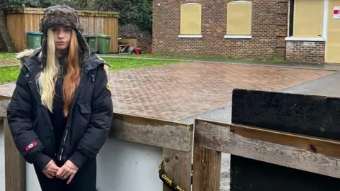 BBC A young woman stands in front of a padlocked gate and boarded up building