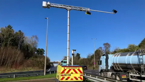 The AI cameras in use which are mounted on a mobile unit painted yellow with red reflective stripes. The cameras are on a raised pole and hang over a road which has a sky blue tanker lorry passing by