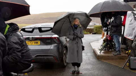 BBC/Silverprint Films/Jamie Simpson TV crew members work around Ashley Jensen, all with umbrellas and waterproof jackets on. Cameras have covers on to protect them from the rain. In the centre of the image, Jensen stands on a marker beside a car, a wet desolate hill behind her, but she is smiling at the camera holding a big golf umbrella, while being wrapped up in a long grey coat, a black beret and a blue and black scarf