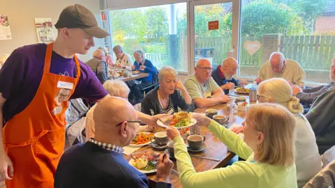 A man in a purple t-shirt, orange apron and black baseball cap is handing a full roast dinner to a woman in a lime green cardigan. She is sat at a cafe table with nine other men and women about to eat the same meal. There is another table of four people in the background eating roast dinners. The cafe has large windows and a glas door at the front.