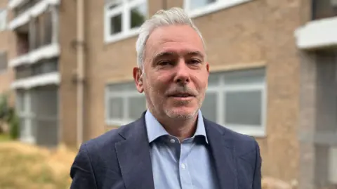 Tom Jackson/BBC Head and shoulders image of Ben Binns. He has grey hair and is wearing a navy suit and light blue shirt, and standing in front of a council block of flats.