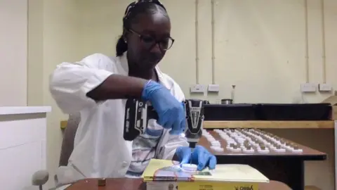 Shamika Spencer, berkacamata, jas lab putih, dan sarung tangan biru, sedang memegang bor di laboratorium tempat dia melakukan eksperimen. Di latar belakang, sebuah meja besar berisi sampel.