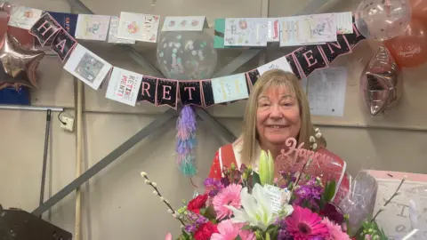 BBC A woman with long blonde hair smiles holding a massive bouquet of flowers. Behind her is black and pink bunting that reads "happy retirement" with several cards hanging up. 