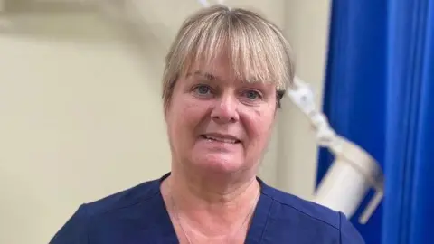 Dr Frances O'Hagan has blonde hair with a full fringe, with her hair tied back. She is smiling at the camera and is wearing navy scrubs. Behind her is an examination bed and a blue privacy curtain but they are both blurred slightly.