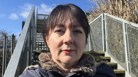 Lara King A dark-haired woman is standing on the steps of a metal footbridge, she is wearing a blue coat with a fur collar and the sky behind her is bright blue with white clouds. 