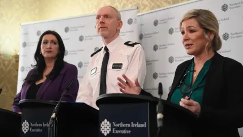 PA Deputy First Minister Emma Little-Pengelly in a purple suit jacket and black top, Ass Ch Cons Davy Beck in PSNI uniform, white shirt with police badges and black tie, and First Minister Michelle O'Neill wearing a black blazer and green blouse, stood at a NI exuctive backdrop with black podiums and microphones as they held a press conference on Thursday at Stormont