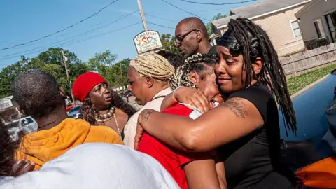 A group of people stand near the site of a mass shooting in Shreveport, Louisiana. One woman, visibly distressed, is comforted by another