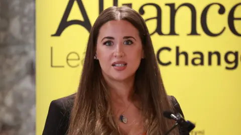 Sorcha Eastwood speaking into a microphone. She has long brown hair and is wearing a silver and blue necklace and a black blazer. 