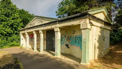 Historic England A one-storey building with six columns at its front which is covered in graffiti. It is surrounded by trees. 
