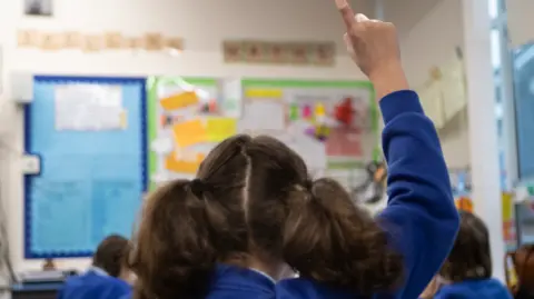 The back of a schoolgirl wearing a blue uniform putting her hand up as she looks towards the front of a classroom.