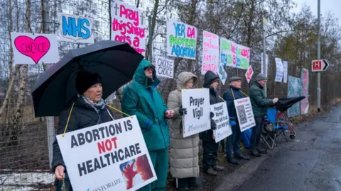 PA Media A group of protestors near an abortion clinic - there are six people, holding placards saying things like Abortion is not healthcare , while also holding umbrellas to shield themselves from the rain. Behind them are a variety of signs pinned to a chain-link fence supporting abortion, saying things like Choose inside a heart.
