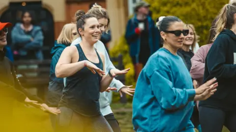 OneRetreat Wellness A woman with her hair tied back in a bun is wearing a blue tank top and moving her hands in the air. She is next to other women who are also dancing outside.