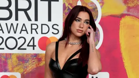A woman with long dark hair and wearing a black dress stands in front of Brit Awards hoarding.