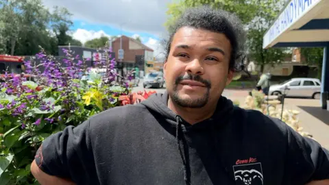 Elliott Smith has black hair and a goatee beard. He is wearing a black hoodie and is stood next to some purple flowers.