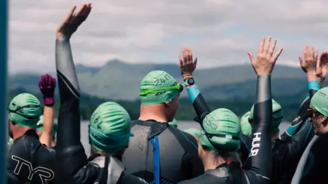 Swimmers wearing wetsuits and tight green swimming hats line up, some with a hand in the air, along the edge of Windermere. On the far side of the lake are the hills of the Lake District 