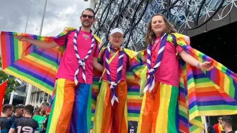 Three people from the Scouts wearing rainbow-inspired outfits stand outside Birmingham's library building