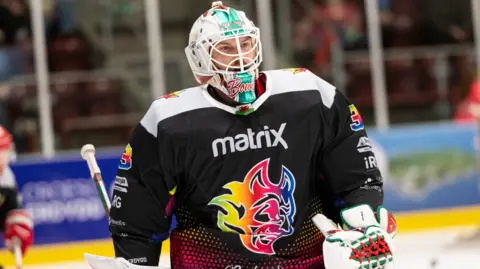 James Assinder/Cardiff Devils Cardiff Devils ice hockey player Ben Bowns in full kit on the ice - his Cardiff Devils logo on his jersey is in the colours of the rainbow as it is a Pride match and he is wearing the gloves and facemask of a goaltender. He is on the ice in an arena and the advertising hoardings can be seen behind him