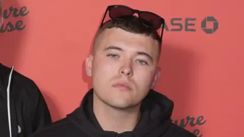 Close shot of a dark haired young man with short brown hair, a black hooded jacket and dark sunglasses on top his head