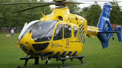 JMG Press An air ambulance which is yellow and blue has landed on grass in a park and a sign on the side says North West Air Ambulance charity