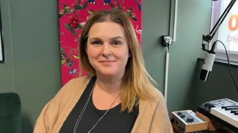 Vikki Irwin/BBC Lucinda Cooper is sitting at a recording desk you can see a microphone and keyboard in the right hand corner of the picture. There is a pink patterned picture with birds and butterflies on the wall behind where Lucinda is sitting. 