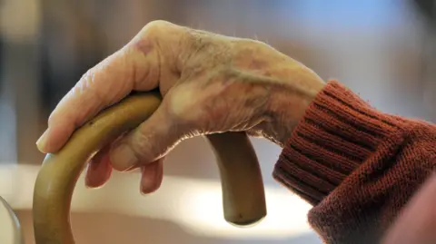 An elderly hand leans on a wooden walking stick, sat down.